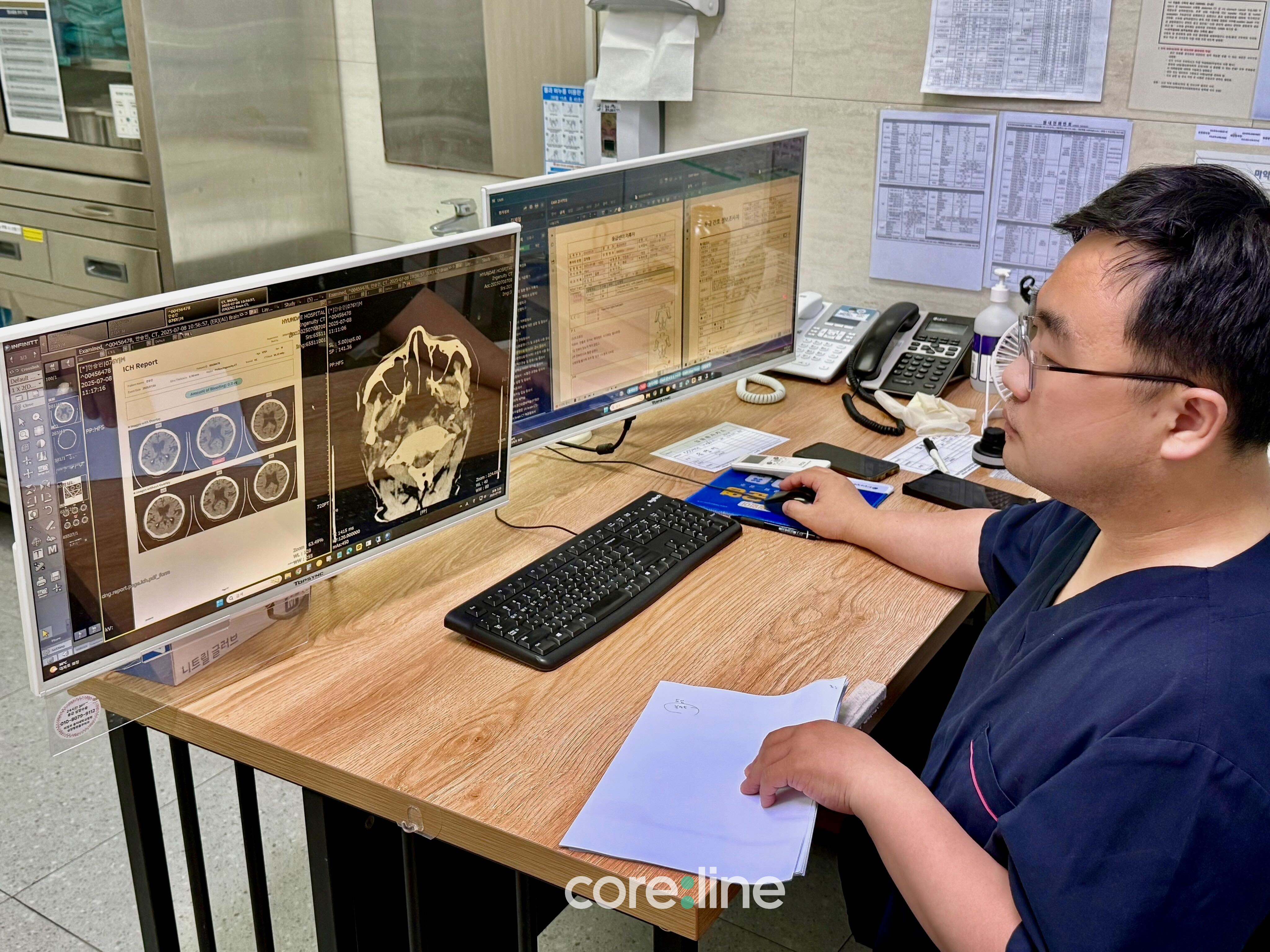 In a hospital consultation room, a clinician reviews CT scans and an AI-generated ICH report on dual monitors. The left screen displays brain CT images with the automated bleeding report, while the right screen shows patient records. The clinician is using a keyboard and mouse while holding patient charts on the desk.